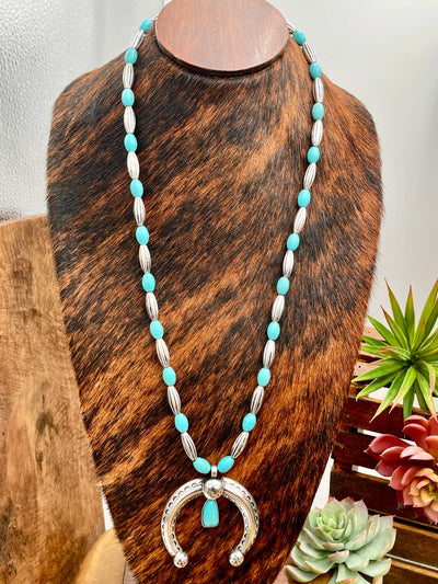 Necklace with turquoise and silver pendant on a wooden stand with plants in the background