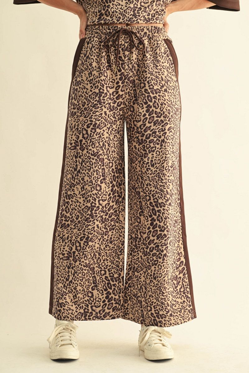A person wearing wide-leg leopard-print pants with a dark brown side stripe, paired with white sneakers, shown from the waist down against a plain light beige studio background.