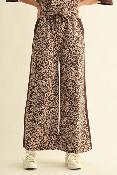 A person wearing wide-leg leopard-print pants with a dark brown side stripe, paired with white sneakers, shown from the waist down against a plain light beige studio background.