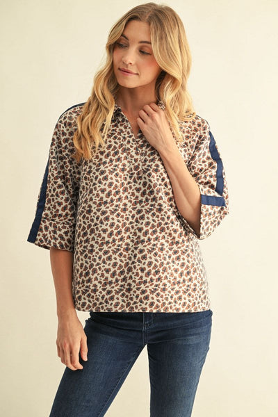 A woman modeling a short-sleeve leopard print blouse with navy blue trim on the sleeves, paired with dark blue jeans, against a plain beige background.