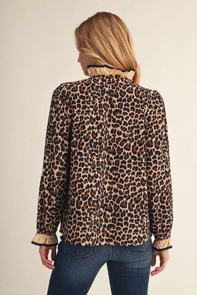 A woman viewed from the back wearing a leopard-print long-sleeve blouse with beige ruffled trim at the collar and cuffs, paired with blue jeans, against a plain light beige studio background.
