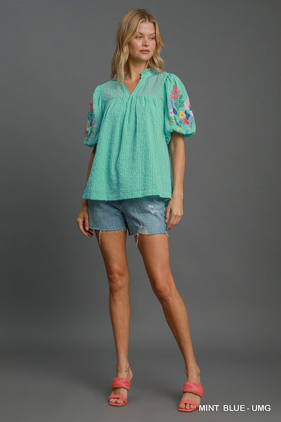 Woman wearing a mint blue blouse with floral embroidery and denim shorts on a gray background