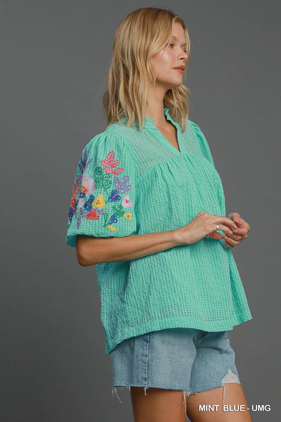 Woman wearing a teal blouse with floral embroidery on a gray background