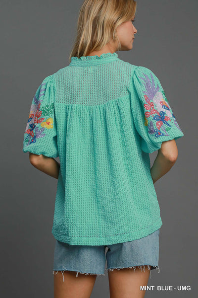 Woman wearing a teal blouse with floral embroidery on a gray background