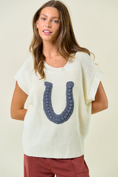 Woman wearing a white knit top with a blue horseshoe design on a plain background