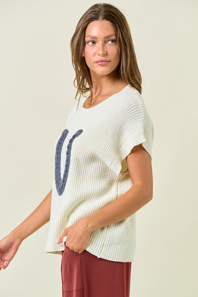 Woman wearing a white knit top with a blue design on a plain background
