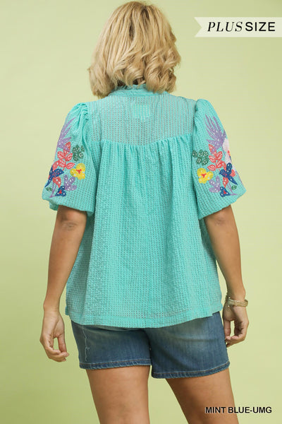 Woman wearing a teal blouse with floral embroidery on a light green background