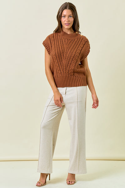 Woman wearing a brown knitted sleeveless top and white pants on a beige background