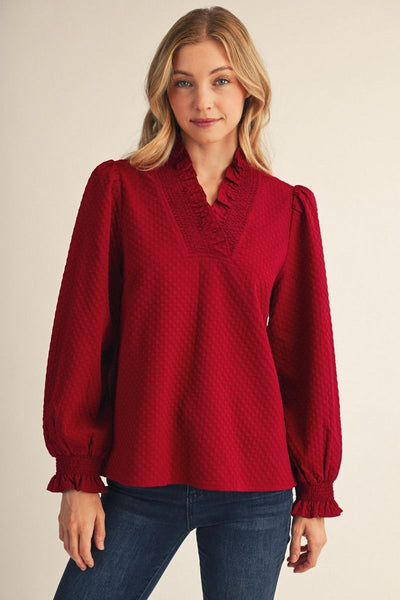 A woman wearing a textured deep red long-sleeve blouse with a ruffled V-neckline and puff sleeves, paired with blue jeans, against a plain beige background.
