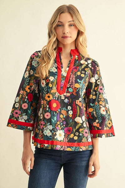 A woman modeling a bohemian-style floral blouse with three-quarter bell sleeves, featuring a multicolor floral print on a dark background with vibrant red trim on the collar, placket, cuffs, and hem, paired with blue jeans against a warm beige studio background.