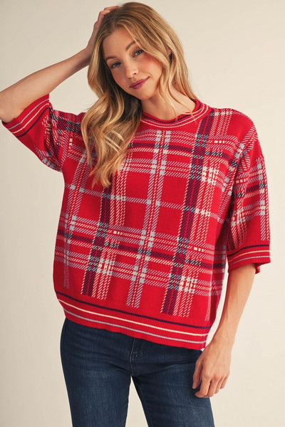 A woman wearing a red plaid knit sweater with white and navy stripes, paired with dark blue jeans, against a plain beige background.