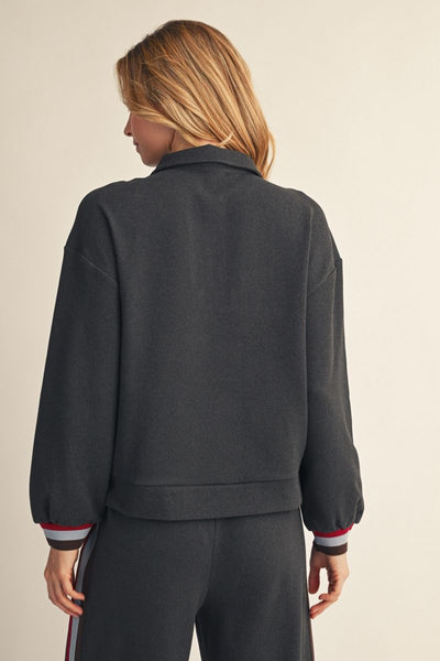 A woman shown from the back wearing a dark charcoal gray cropped jacket with ribbed cuffs featuring red, white, and navy stripes, paired with matching dark gray pants against a light beige background.