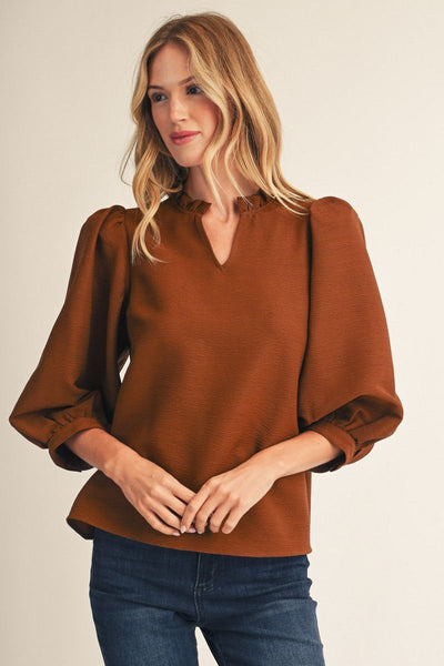 A woman wearing a rust-brown long-sleeve blouse with a split V-neckline and gathered sleeves, paired with dark blue jeans, against a plain light beige background.