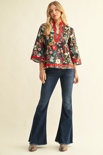 A woman modeling a bohemian-style floral blouse with three-quarter bell sleeves, featuring a multicolor floral print on a dark background with vibrant red trim on the collar, placket, cuffs, and hem, paired with blue jeans against a warm beige studio background.