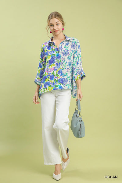 Woman wearing a floral blouse and white pants, holding a blue handbag on a light green background.