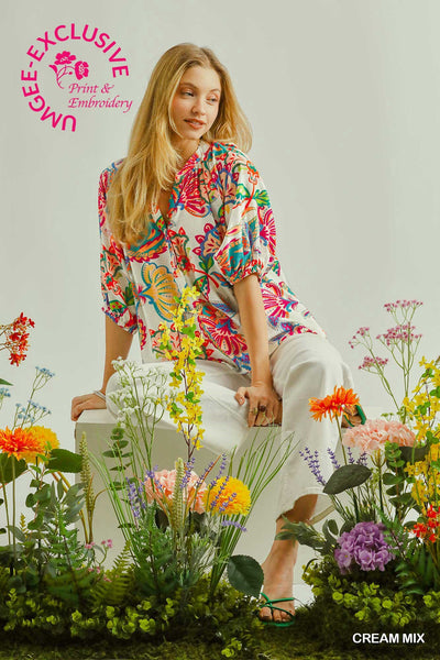 Woman in a colorful floral dress sitting among flowers with Umgee Exclusive branding.