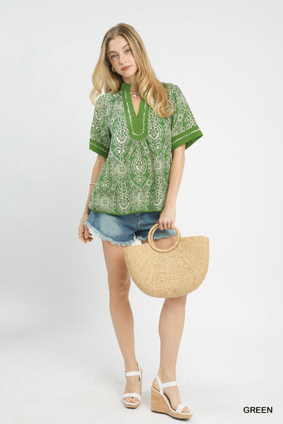 Woman wearing a green patterned blouse with denim shorts, holding a straw bag on a white background.