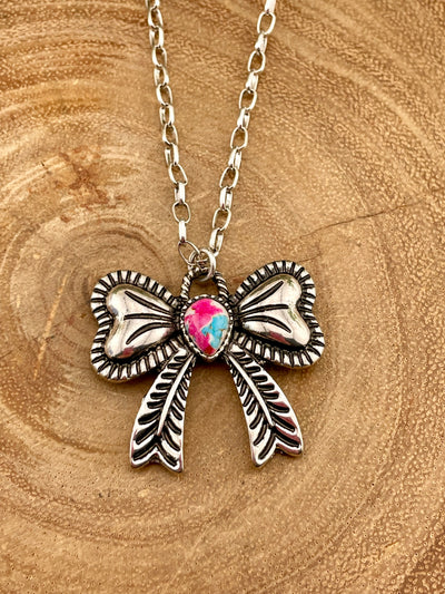 Bow necklace on a wooden surface with a pink & turquoise mix center stone.