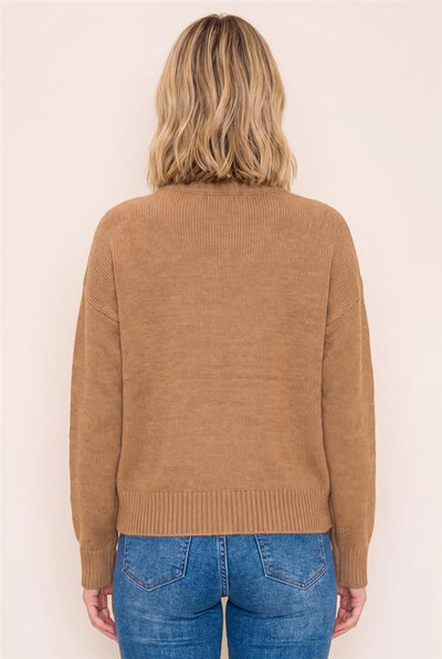 A woman viewed from the back wearing a camel brown ribbed knit sweater and blue jeans against a plain off-white background.