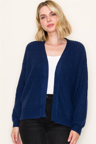 Woman wearing a blue cardigan over a white top and black pants on a plain background