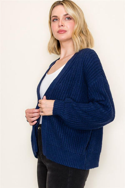 Woman wearing a blue cardigan over a white top and black pants on a light background