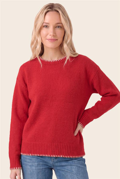 A woman wearing a red knit sweater with white decorative trim around the crew neck, cuffs, and hem, paired with blue jeans, against a solid light beige background.