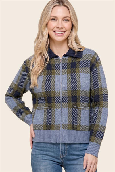 A woman wearing a zip-up plaid sweater with a navy collar, blue and olive green check pattern, and ribbed cuffs and hem, paired with blue jeans, against a solid beige background.