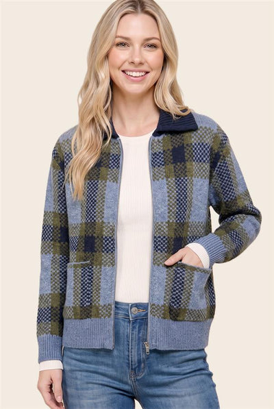 A woman wearing a blue and green plaid zip-up cardigan over a white ribbed top and blue jeans, photographed against a plain light beige studio background.