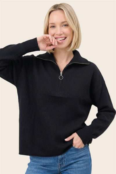 A woman wearing a black long-sleeve quarter-zip sweater with a silver zipper pull, paired with blue jeans, against a solid light beige background.