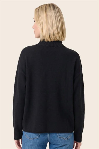 A woman with blonde hair is shown from the back wearing a black mock neck sweater and blue jeans against a solid light beige background.
