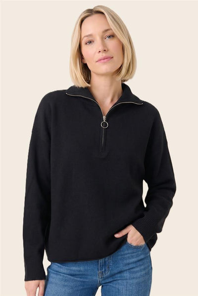 A woman with a short blonde bob wearing a black long-sleeve quarter-zip sweater with a silver-tone ring pull, paired with blue jeans, against a solid light beige background.