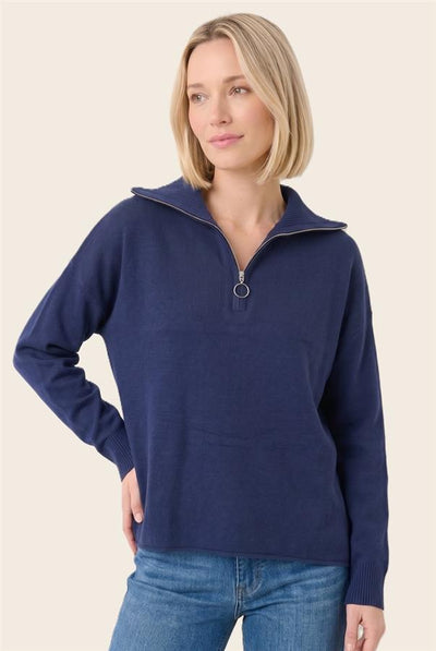 A woman modeling a navy blue half-zip pullover sweater with a ribbed collar and cuffs, paired with blue jeans, against a solid light beige background.
