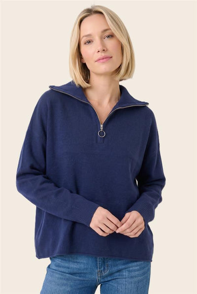 A woman wearing a navy blue half-zip mock-neck sweater with a silver zipper pull, paired with blue jeans, against a solid light beige background.