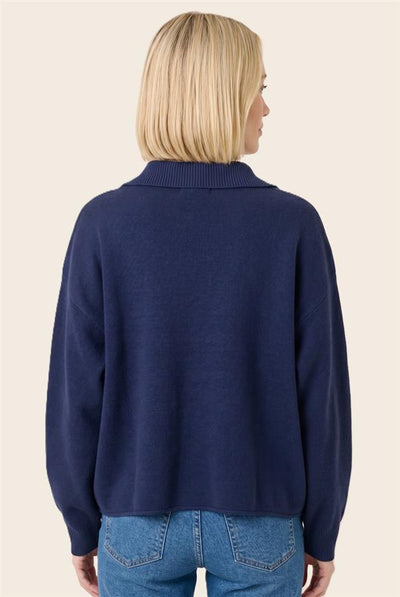 A woman with blonde hair is shown from the back wearing a navy blue knit sweater and blue jeans against a solid light beige background.