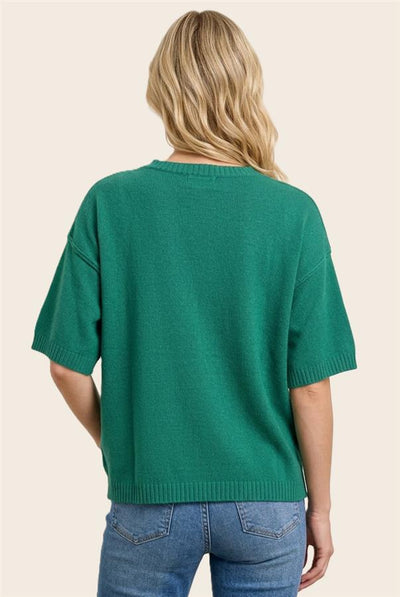 A woman viewed from the back wearing a green short-sleeve knit sweater and blue jeans against a plain light beige background.