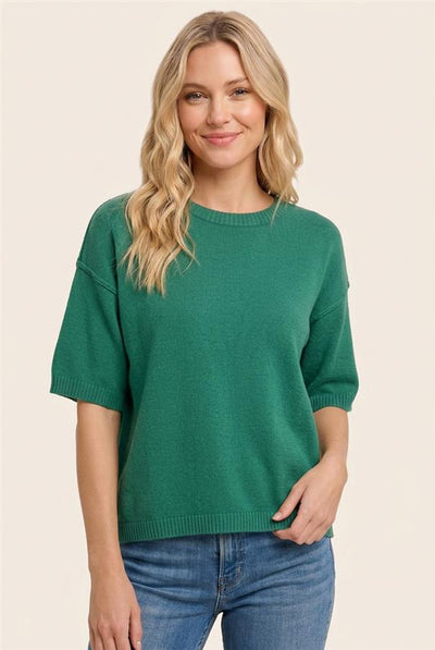 A woman wearing a green short-sleeve knit sweater and blue jeans, standing against a plain light beige background.