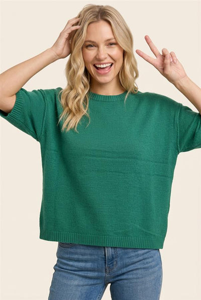 A woman wearing a green short-sleeve knit sweater and blue jeans, posing with her hands raised against a solid light beige background.