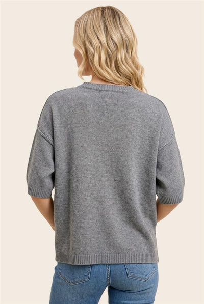 A woman viewed from the back wearing a heather gray short-sleeve knit sweater and blue jeans against a plain light beige studio background.