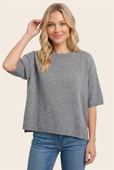 A woman wearing a heather gray short-sleeve knit sweater and blue jeans, standing against a solid light beige background.