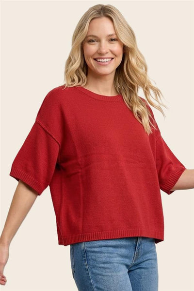 A smiling blonde woman wearing an oversized short-sleeve knit sweater in a rich red color, paired with blue jeans, against a solid light beige background.