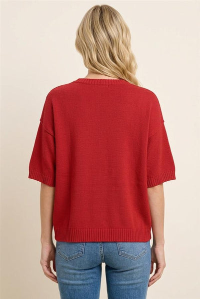 A woman viewed from the back wearing a red knit short-sleeve sweater and blue jeans against a plain beige studio background.