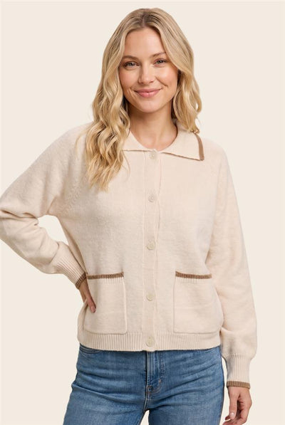 A woman wearing a cream-colored knit cardigan sweater with brown trim details, paired with blue jeans, against a solid light beige background.