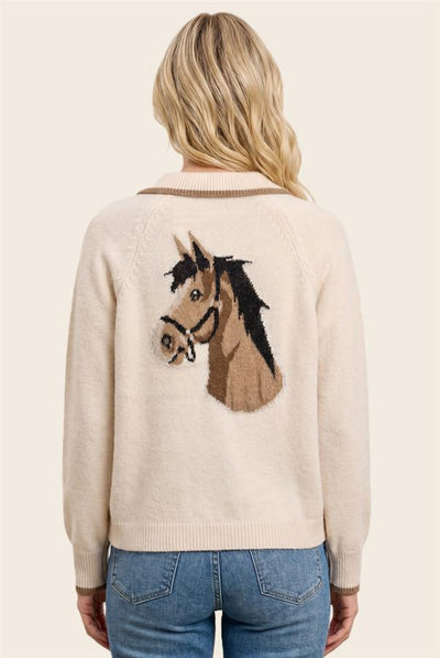 A woman viewed from the back wearing a cream-colored knit sweater with a large embroidered horse head design on the back, paired with blue jeans, against a solid light beige background.