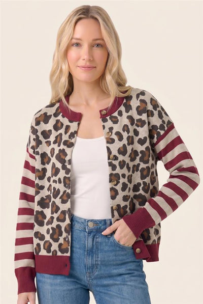 A woman modeling a leopard-print and striped knit cardigan with burgundy ribbed trim, worn over a white tank top and blue jeans against a plain beige studio background.