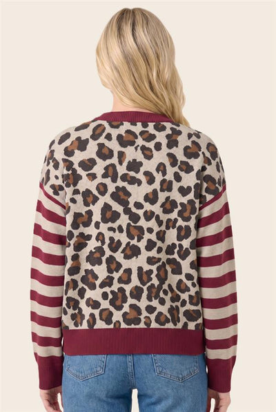 A woman viewed from the back wearing a knit sweater with a leopard print body and burgundy-and-cream striped sleeves, paired with blue jeans, against a plain light beige background.