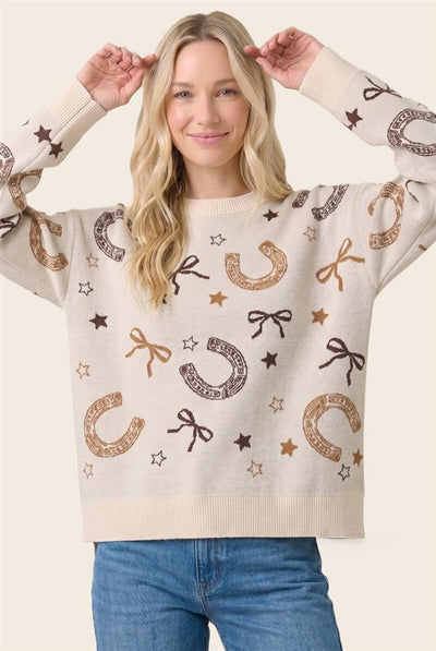 A woman wearing a cream-colored knit sweater with a western-themed pattern of brown and gold horseshoes, bows, stars, and the word 'RANCH'. She is paired with blue jeans, posing against a solid light beige background.