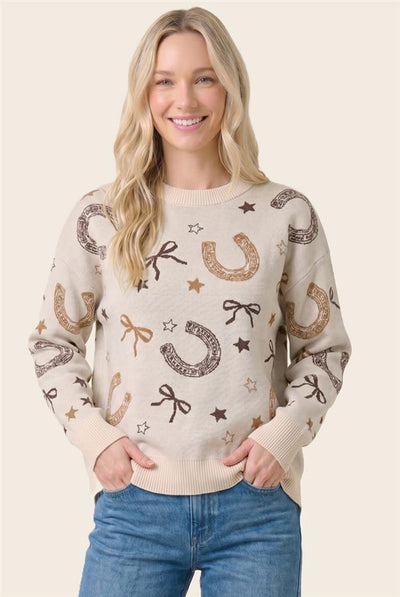 A woman wearing a cream-colored knit sweater with a western-themed pattern of brown and gold horseshoes, bows, stars, and the word 'RANCH'. She is paired with blue jeans, posing against a solid light beige background.