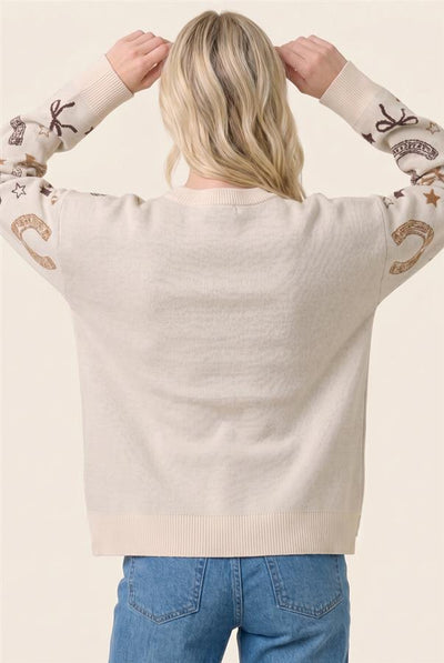 A woman viewed from behind wearing a cream-colored knit sweater with western-themed embroidery on the sleeves, paired with blue jeans, against a solid light beige background.