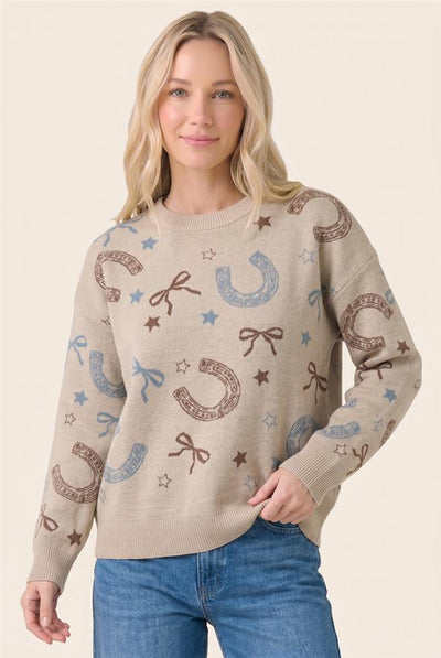 A woman wearing a beige crewneck sweater with an allover pattern of brown horseshoes, blue bows, and blue stars, paired with blue jeans, against a solid light beige background.