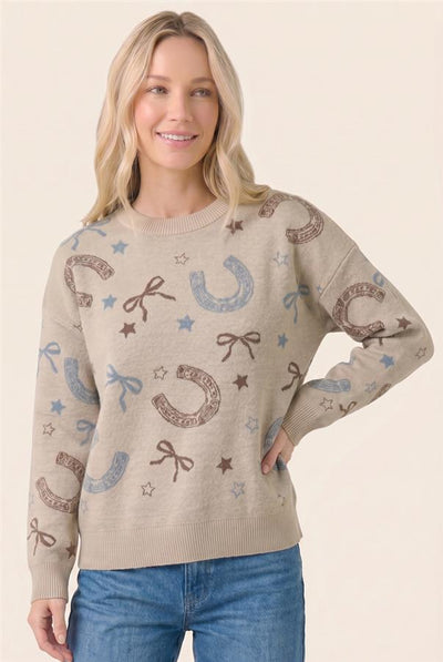 A woman wearing a beige crewneck sweater with an allover pattern of brown horseshoes, blue bows, and blue stars, paired with blue jeans, against a solid light beige background.
